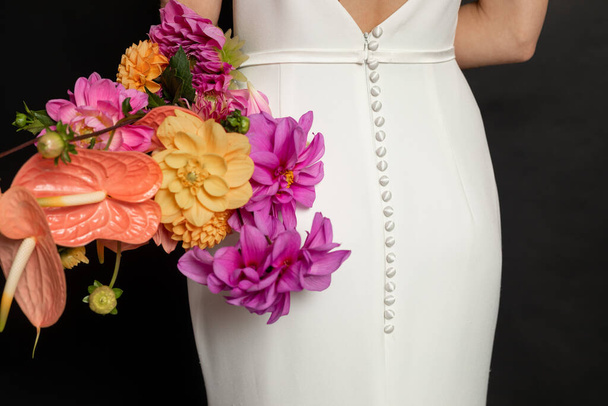 ボタン付きの白いドレスでエレガントな女性の認識できないクロップバックビューは、黒の背景にカラフルな花の花束を保持します。結婚式と結婚式婚約。休日の招待状。モックアップ - 写真・画像