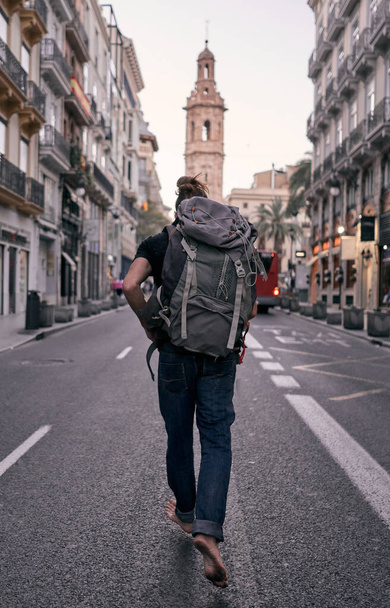 young caucasian man running along the asphalt of a city street with a backpack on his back barefoot with dirty feet, valencia, spain - Backpacker travel concept - Zdjęcie, obraz