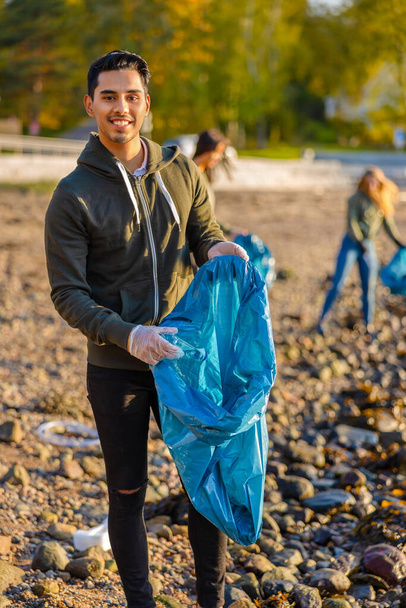 Full length portrait of young man picking up garbage in bag. Multi-ethnic group of volunteers are cleaning beach against sky. They are protecting environmental from damage. - Photo, image