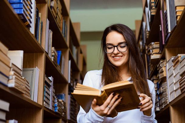 Cheerful student-girl, standing between bookshelves and reading a book. Express positive emotions while reading, intellectual leisure time. - Fotografie, Obrázek