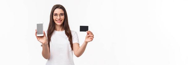 Iloisen ystävällisen näköisen nuoren naisen muotokuva t-paidassa ja laseissa, jossa näkyy luottokortti ja matkapuhelinnäyttö mainoksena online-sovellus, internet-osto, muu kuin käteismaksu. - Valokuva, kuva