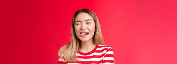 Közelkép pimasz pimasz gondtalan kimenő városi ázsiai nő diák élvezni nyári nyaralás viselni csíkos póló, show nyelv játékosan kacsintó kamera, állni piros háttér pozitív barátságos hangulat. - Fotó, kép