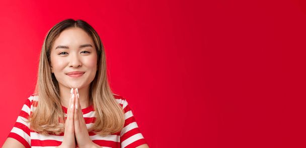 Thankful cute happy asian girl bowing politely show namaste gesture gladful. Joyful attractive blonde hold hands pray appreciate help begging favour, stand relaxed outgoing red background. - Photo, Image