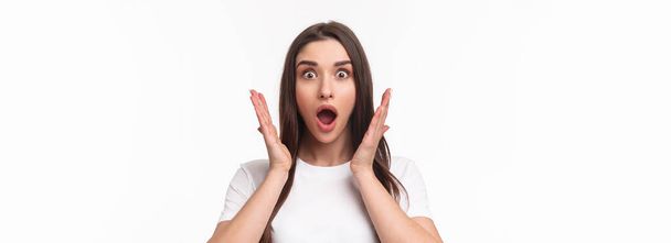 What an amazing news. Surprised and astonished, excited young woman react to something awesome happened, gasping, open mouth and raise hands near face, staring camera, white background. - Photo, Image