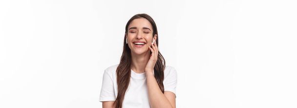 Portrait en gros plan d'une femme caucasienne adorable, souriante et riante les yeux fermés, touchant des écouteurs sans fil, ayant une conversation téléphonique avec des écouteurs, fond blanc. - Photo, image