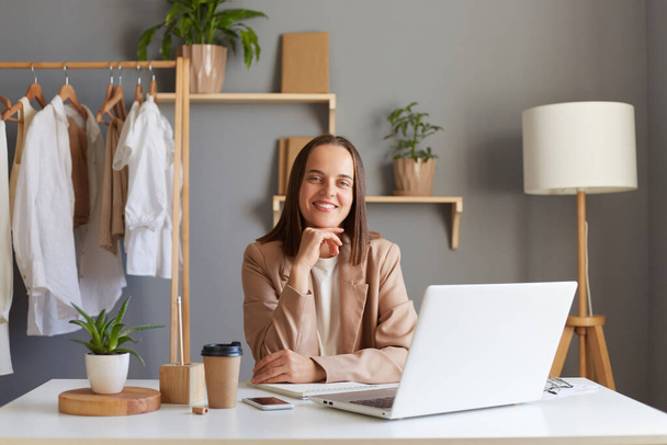 Muotokuva hymyilevä onnellinen tyytyväinen ruskeahiuksinen nainen yllään beige takki istuu työpaikallaan edessä kannettavan, katsoen kameraa, pitäen leuka, ilmaista positiivisia tunteita työssään. - Valokuva, kuva