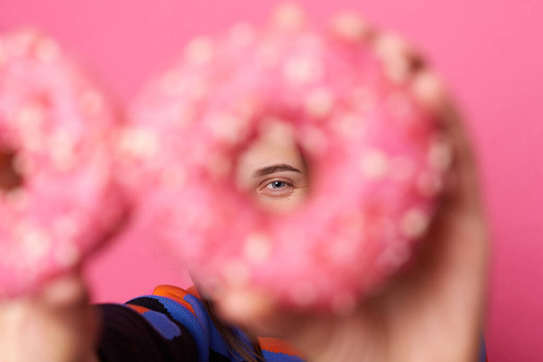 Portrait of woman holding in hands and looking though tasty doughnut with bright sprinkles at camera with cheerful expression, isolated over pink background. - Photo, Image