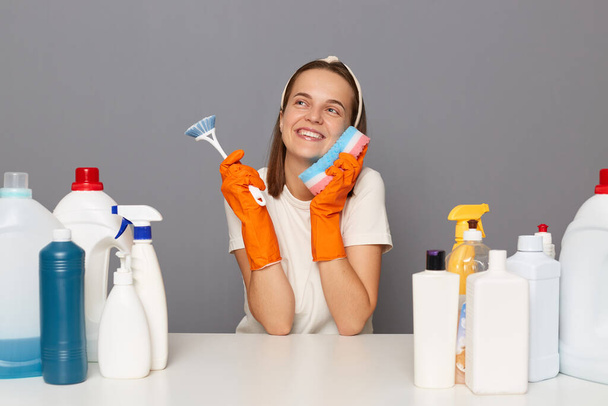 Indoor shot of smiling positive happy Caucasian woman wears headband, gloves and casual t shirt, posing isolated over gray background, smiles broadly, dreaming, surrounded with bottles of detergents. - Φωτογραφία, εικόνα