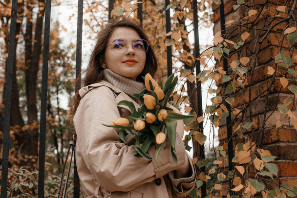 Portrait of young stylish attractive woman walking in city, street fashion, autumn trend, coat, holding flowers bouquet, elegant style, yellow tulips, romantic mood. Outdoor portrait of young girl. - Foto, Imagen