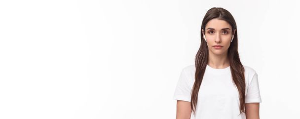 Close-up portrait of serious-looking attractive caucasian woman in wireless earphones, looking at camera with relaxed no emotions face, standing white background. Copy space - Photo, Image