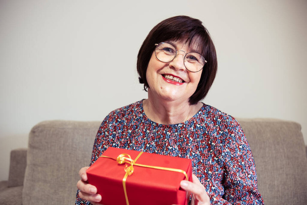 Une femme âgée souriante avec des lunettes dans une robe florale tenant présent dans une boîte rouge avec un ruban d'or dans ses mains - Photo, image
