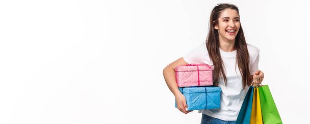 Shopping, holidays and lifestyle concept. Portrait of happy pleased young girl likes making presents and celebrating, holding wrapped bags of gists, laughing and walking to friend birthday. - Photo, Image
