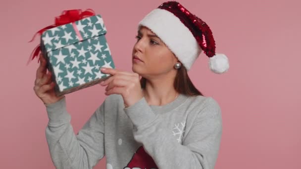 Запутавшаяся любопытная молодая женщина в свитере Санта-Клауса празднует рождественские праздники встряхивая завернутую рождественскую подарочную коробку доставки, держа возле уха, слушая угадать, что внутри. Девушка на розовом фоне - Кадры, видео