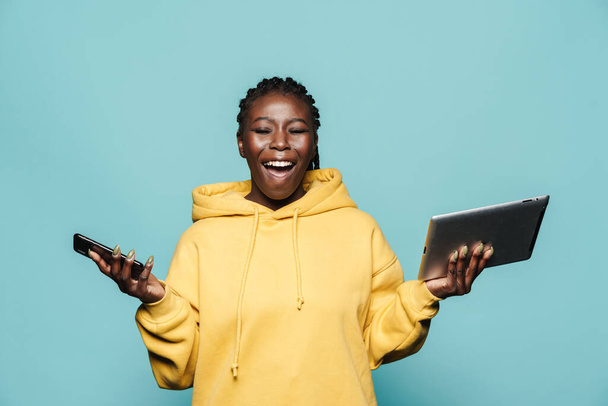 Young black woman laughing while posing with gadgets isolated over blue background - Fotografie, Obrázek