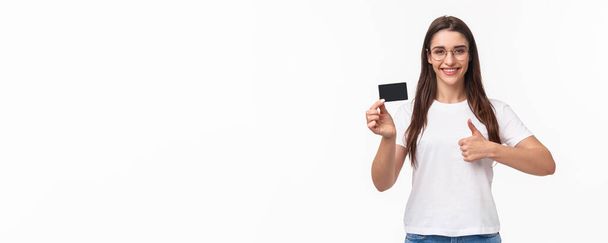 Portret zadowolonej, atrakcyjnej młodej brunetki w t-shircie i okularach, polecam bank, pokazujący kartę kredytową, uśmiechnięty zadowolony pokazać kciuki do góry, aprobuję i doradzam dokonywać zakupów online. - Zdjęcie, obraz