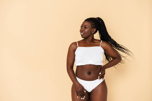 Black young woman with afro pigtails smiling while posing in underwear isolated over beige background - Foto, imagen