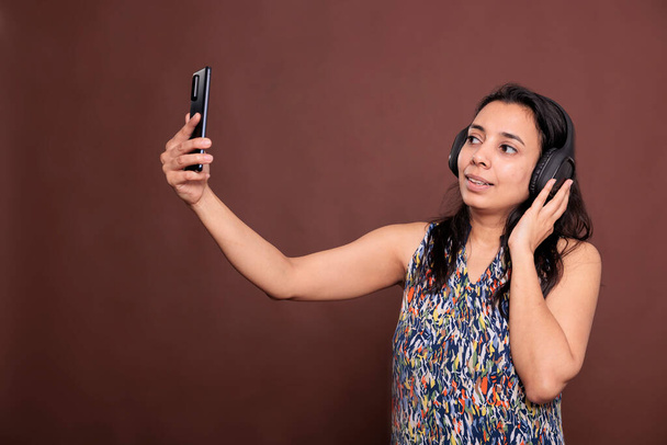 Hinduska kobieta w słuchawkach rozmawiająca przez smartfona na spotkaniu online, rozmawiająca przez telekonferencję, patrząca na kamerę telefonu komórkowego. Rozmowa wideo, komunikacja zdalna, wideokonferencja - Zdjęcie, obraz