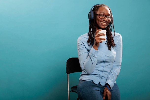 Cheerful happy african american woman smiling heartily while listening to music playlist on wireless headphones. Joyful positive young adult person with earphones enjoying radio on blue background. - Foto, afbeelding