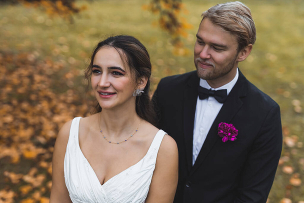 Romantic autumn wedding photoshoot. Dreamy positive beautiful Middle Eastern woman in sleeveless wedding gown standing in front of her Scandinavian blond husband. Medium closeup outdoor shot. High - Photo, Image