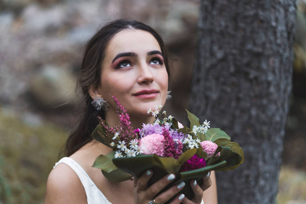 Bridal photoshoot concept. Beautiful ethnic bride posing outdoor in the forest. Closeup portrait of attractive Turkish woman looking up and holding her bridal bouquet next to her face. High quality - Fotoğraf, Görsel
