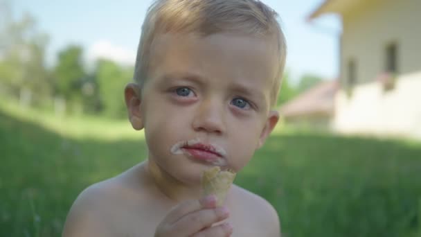 Aranyos kisfiú fagylaltkúpot eszik a kert árnyékában egy nyári napon. Imádnivaló kisbaba, vaníliás fagyit eszik a hátsó udvar árnyékos részén. Teljes felfrissülés egy forró nyári napon. - Felvétel, videó