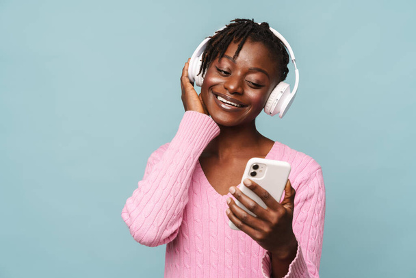 African american young woman smiling while using cellphone and headphones isolated over blue background - Фото, изображение