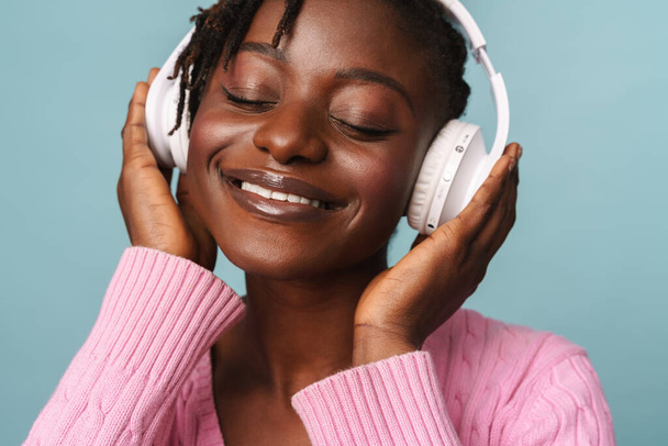 African american young woman smiling while listening to music with headphones isolated over blue background - Фото, зображення