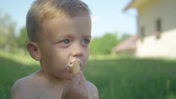 閉じる:暑い日に庭の影でアイスクリームを楽しむ愛らしい男の子。裏庭の日陰の部分でバニラアイスコーンを楽しんで甘い幼児。暑い夏の日の完全なリフレッシュ. - 映像、動画