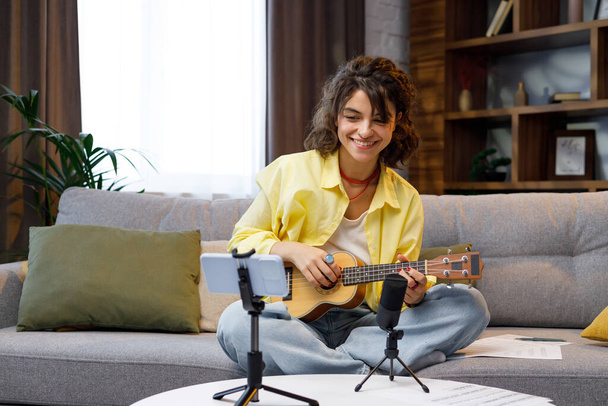 Blogger with mobile phone and microphone. A beautiful hipster girl in a yellow shirt plays and sings on the ukulele on the sofa in the apartment. Online learning to play the guitar - Foto, afbeelding