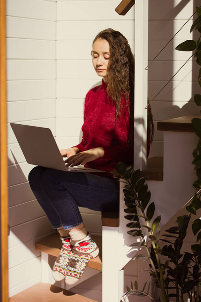 Egy gyönyörű fiatal lány, fiatal nő, aki egy laptopon dolgozik otthonról. Függőleges portré egy meleg ruhás diákról, kötött zokni ül a lépcsőn egy hangulatos otthoni légkörben ősszel, téli napon - Fotó, kép