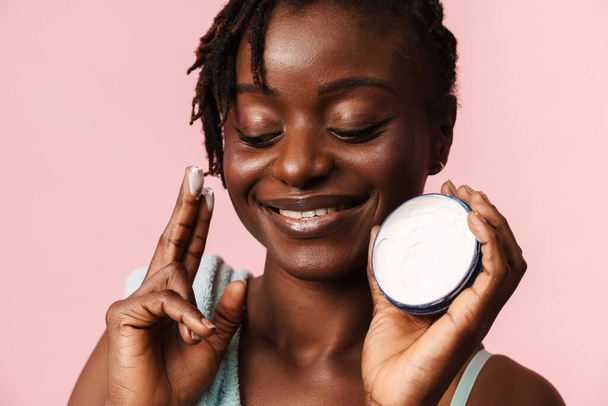 Black shirtless woman with towel smiling while applying face cream isolated over pink background - Fotografie, Obrázek
