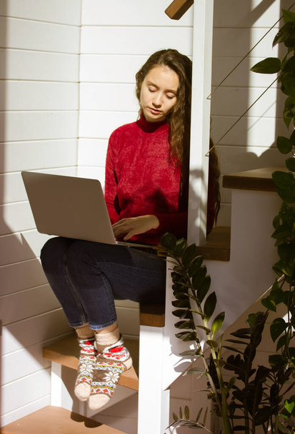 Una hermosa joven, una joven que trabaja en un portátil desde casa. Retrato vertical de un estudiante en ropa de abrigo, calcetines de punto sentado en las escaleras en un ambiente acogedor en el otoño, día de invierno - Foto, imagen