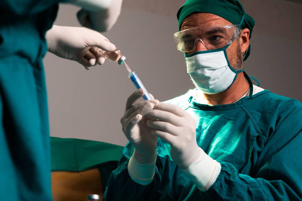 外科医は、補助看護師と共に滅菌手術室で外科手術用バイアルから注射器を埋めます。手術のための完全な保護服の医師や医療スタッフは麻酔注射を準備します - 写真・画像