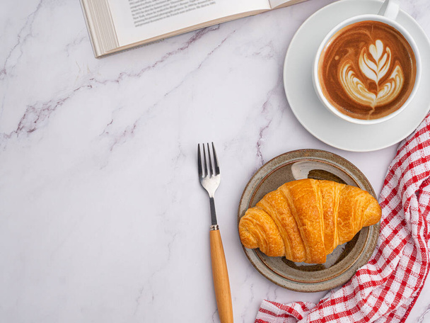 Top view of a book with croissant on a plate and a white coffee cup placed on a marble background. Space for text. Concept of beverage and relaxation - 写真・画像