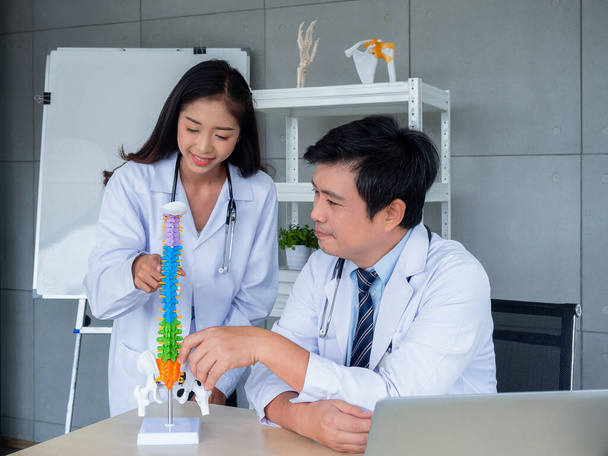 Twee Aziatische artsen in witte uniformen praten en werken samen in het medisch kantoor. De orthopedische volwassen mannelijke arts biedt kennis en begeleiding aan vrouwelijke arts over wervelkolom op bureau in het ziekenhuis. - Foto, afbeelding