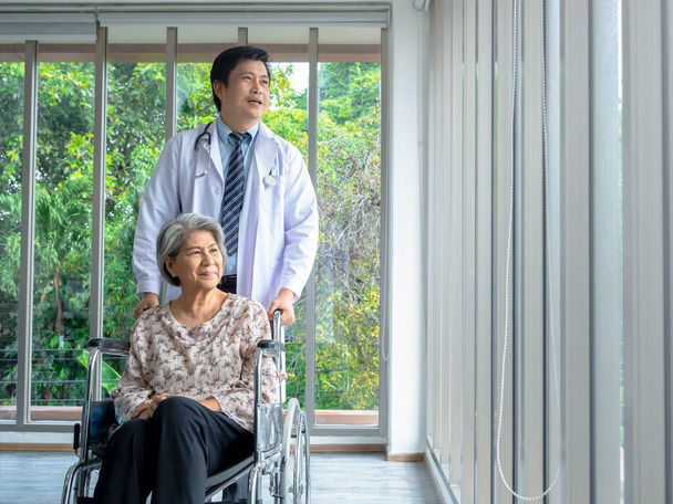 Lachend gelukkig Aziatische oudere oudere vrouwelijke patiënt zit in een rolstoel terwijl de man arts in witte jas staat in het medisch kantoor in het ziekenhuis, verticale stijl. Verzorging, gezondheidszorg en medisch concept. - Foto, afbeelding