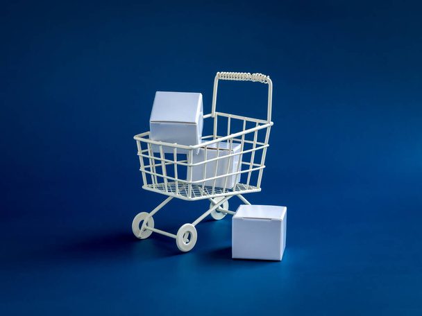 ショッピングオンラインビジネス、電子商取引、配信、購買力の概念。青の背景に隔離されたショッピングトロリーカートのスーパーマーケットで白い小包ボックス、最小限のスタイル. - 写真・画像