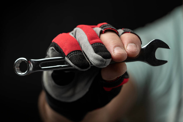 Сильная мужская рука в красной перчатке держит гаечный ключ на черном фоне. Концепция сантехнических, механических и автомобильных работ. - Фото, изображение