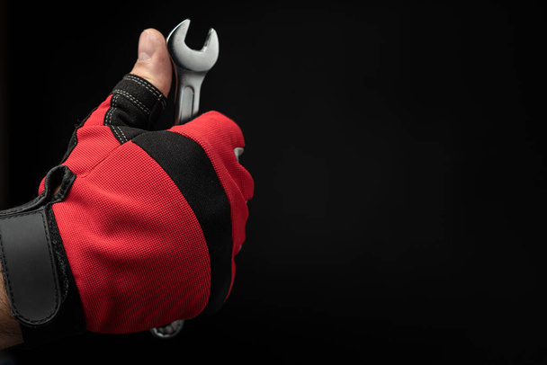 Сильна чоловіча рука в червоній рукавичці тримає ключ на чорному тлі. Концепція сантехніки, механічних і автомобільних робіт
. - Фото, зображення