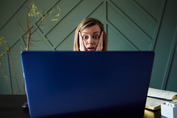 Dizüstü bilgisayara yuvarlak gözlerle bakan şok olmuş bir kadın. Çevrimiçi haberlerde heyecanlı duygusal tepkiler - Fotoğraf, Görsel