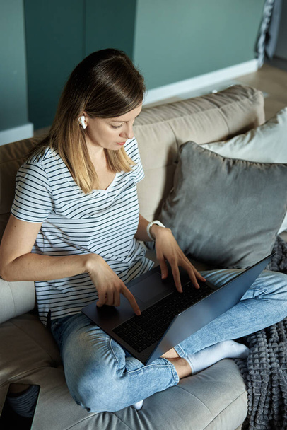 Woman sitting on sofa in living room and using laptop. Female freelancer working at home office. Remote work and work from home concept. - Fotó, kép