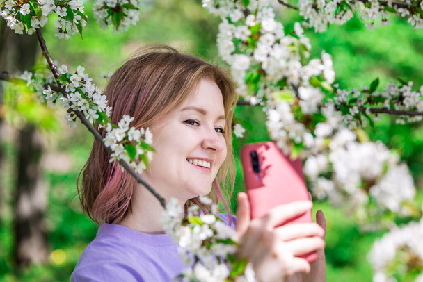 Vrouw in een paars t-shirt die naar haar vrienden gaat op een videochat. Een tedere foto met witte bloemen in de appelboomgaard. Smartphone en technologie concept. - Foto, afbeelding