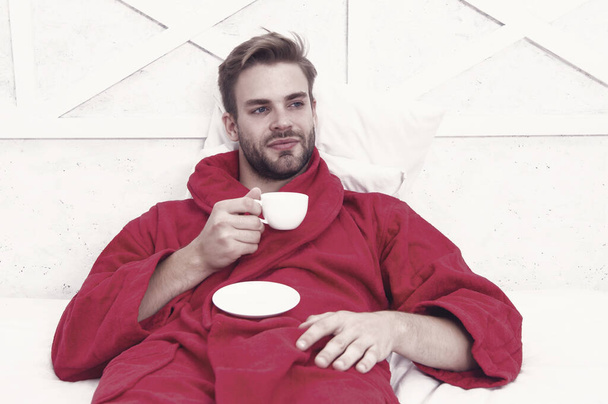 His favorite morning beverage. Handsome guy in bathrobe holding cup of hot drink. Bearded caucasian man enjoying his beverage in bed. Relaxing with tasty beverage. - Fotó, kép