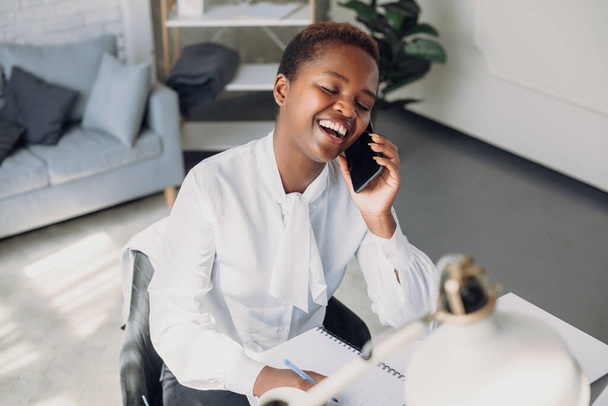 Retrato de una joven mujer negra sonriente usando su teléfono inteligente y su computadora portátil moderna, sentada en el escritorio de la oficina. Comunicación por Internet. Estilo de vida - Foto, imagen