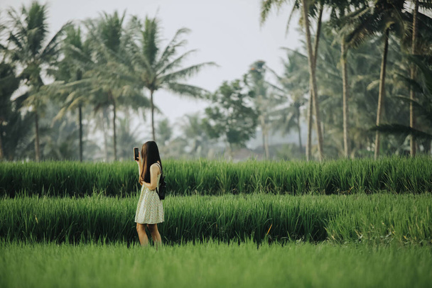 Hi-res photo of an south east asian teenager girl traveling and capture a photo using smartphone in the middle of rice field with blurred background - Fotó, kép