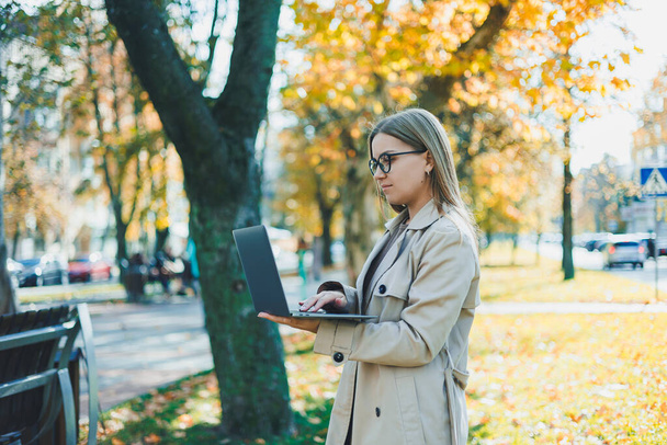 Una donna carina in occhiali con i capelli lunghi sta camminando in un parco autunnale con un computer portatile in mano. Lavora online all'aria aperta - Foto, immagini