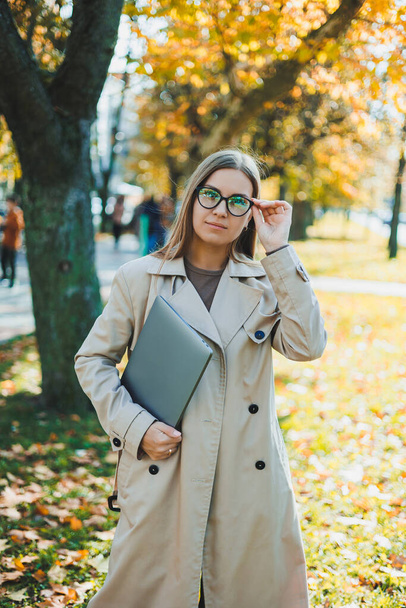 A cute woman in glasses with long hair is walking in an autumn park with a laptop in her hands. Work online in the fresh air - Foto, afbeelding