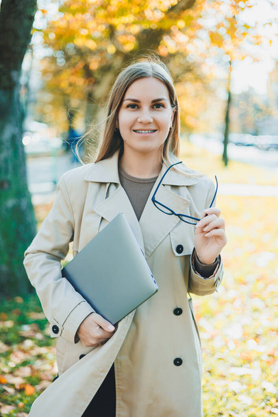 A cute woman in glasses with long hair is walking in an autumn park with a laptop in her hands. Work online in the fresh air - Fotografie, Obrázek