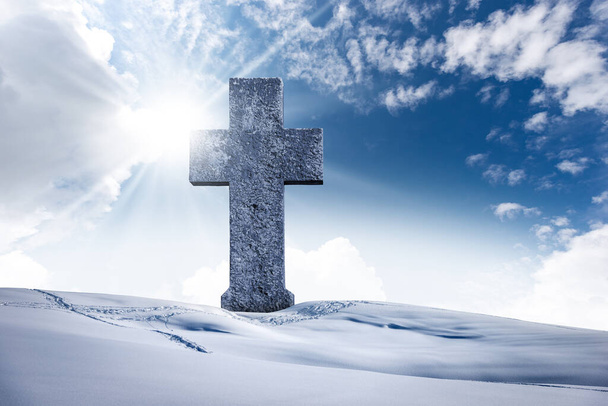 Gran cruz cristiana de piedra en un paisaje de montaña con nieve contra un cielo azul claro con nubes, rayos de sol y espacio de copia. - Foto, imagen