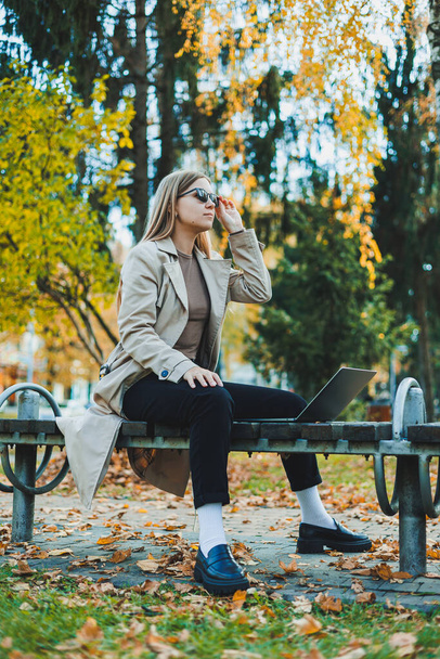 Cute female student sitting in an autumn park with a laptop and working online. Happy woman working outdoors in the park. - Foto, immagini